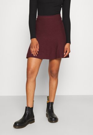 Femme portant une mini-jupe rouge foncé, un haut noir à manches longues et des bottines noires, debout devant un fond clair uni.