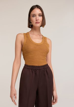 Frau mit kurzen braunen Haaren trägt ein senfgelbes ärmelloses Stricktop und dunkelbraune hoch taillierte Hosen, steht vor einem einfarbigen Hintergrund.