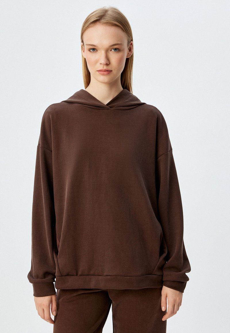 Sudadera marrón con un ajuste relajado, textura suave y un bolsillo tipo canguro. Presenta una capucha con cordón, mangas largas y puños anchos.