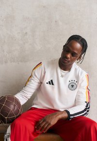 Hvit langermet sweatshirt med svarte og røde detaljer, med en logo og tre stjerner. Røde bukser, holder en brun basketball.