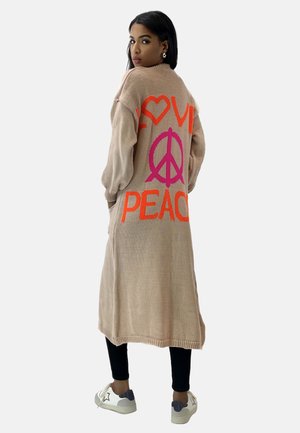 Lange beige cardigan met "LOVE" en "PEACE" in helder oranje en roze, gebreide textuur, geribbelde rand, gedragen over zwarte leggings en sneakers.