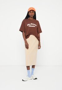 Camiseta castanha com estampado "New Balance Athletics", saia midi bege, meias azul claro, sapatilhas brancas e um boné laranja.