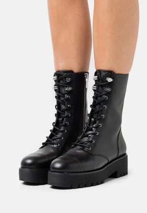 Zwarte leren veterlaarzen met een zijrits en een dikke geribbelde zool, gedragen op blote benen tegen een witte achtergrond.