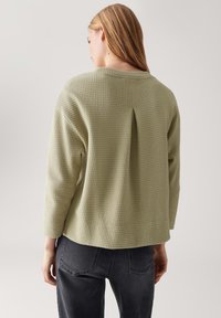 Donna con capelli castano chiaro che indossa una maglia a maniche lunghe verde chiaro a trama e jeans grigio scuro, girata di spalle su uno sfondo semplice.