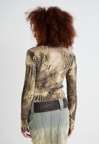 Top à manches longues dans un vert olive atténué et brun, avec un motif texturé. Associé à une jupe en denim dégradée bleu clair et une taille sombre cloutée.