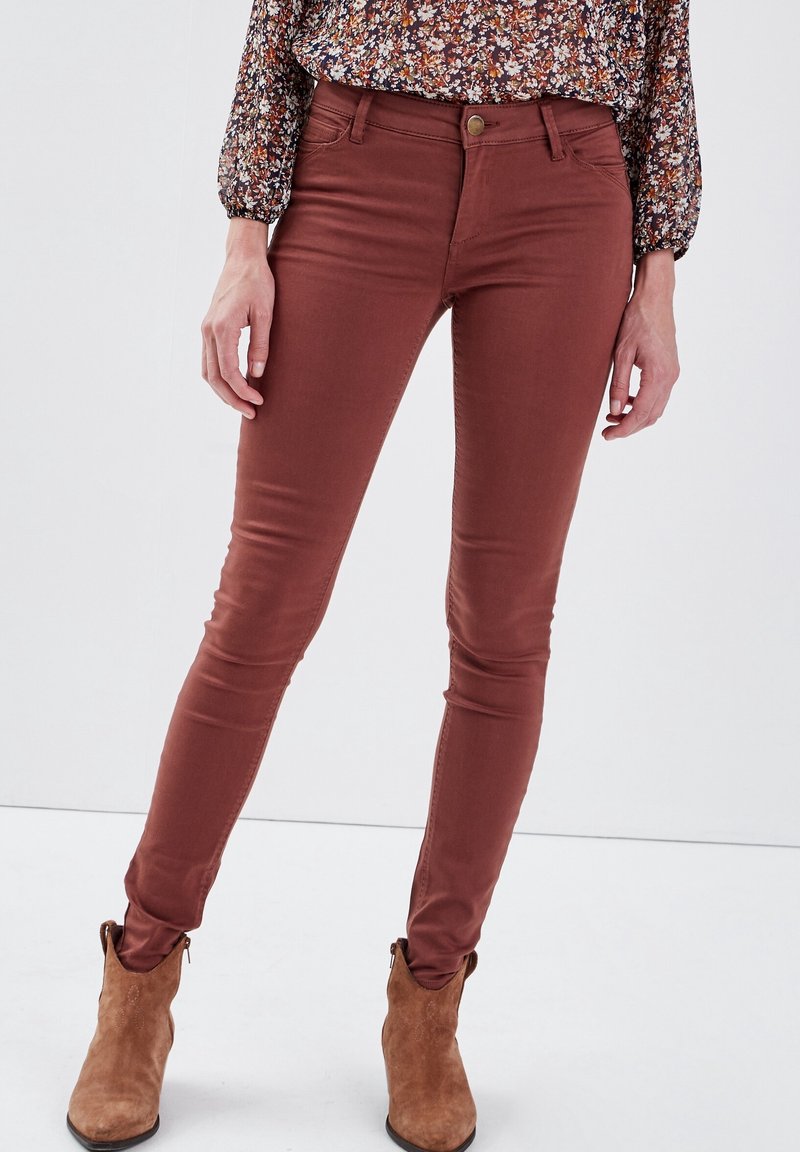 Femme portant un pantalon slim couleur rouille, un chemisier à manches longues à fleurs et des bottines en daim camel, debout devant un fond blanc.