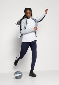 Abbigliamento sportivo composto da una giacca grigia chiara con dettagli scuri, una maglietta bianca e pantaloni blu navy. Scarpe nere; pallone da calcio grigio e bianco.