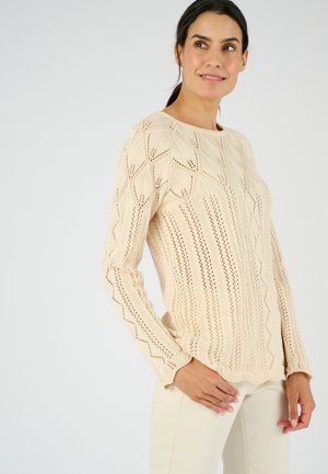 Vrouw met donker haar die een lichtbeige gebreide trui met patroon en crèmekleurige broek draagt, staand met één hand bij haar heup, kijkt naar rechts.