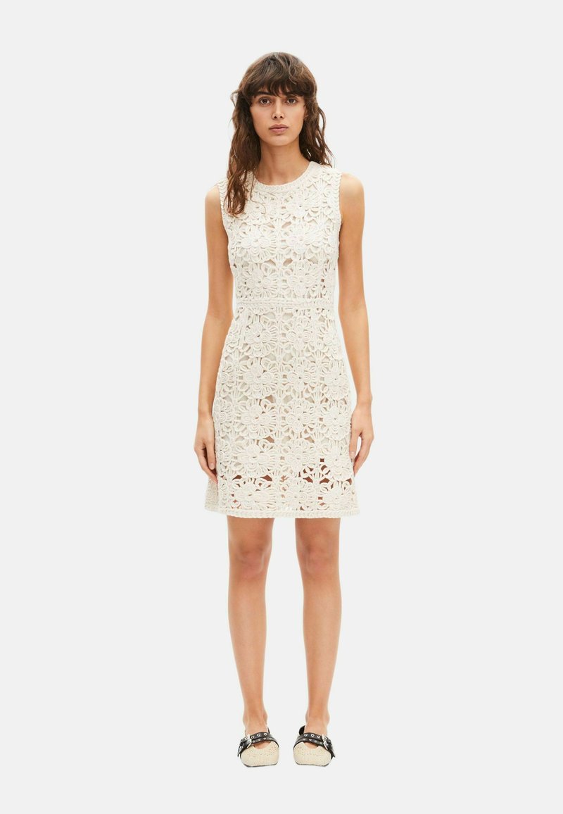Mujer de pie con un vestido blanco sin mangas de encaje floral por encima de la rodilla y zapatos planos blancos y negros, sobre un fondo liso.