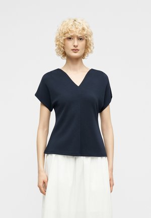 Woman with curly blonde hair wearing a navy blue V-neck short-sleeve top and a white skirt, standing against a plain light background.