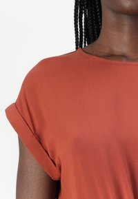 Top v rezavé barvě s krátkým rukávem a volným střihem, s ohrnutými manžetami. Hladká látka s lehkou texturou, vhodná pro každodenní nošení.