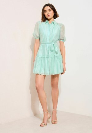 Naine kannab helerohelist, lühikeste varrukatega nööbitavat kleiti vöökohal paelaga ja kuldseid kõrge kontsaga sandaale, seisab ühtlase hele taustaga ees.