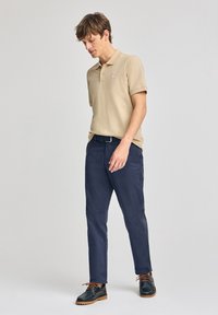 Beige kortärmad polo skjorta med en liten logotyp, ihopparad med marinblå chinos och mörka snörskor med bruna accenter.