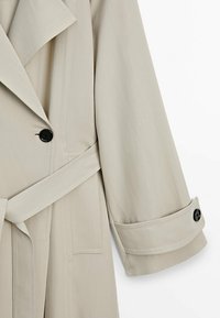 Beżowy płaszcz trench wykonany z tkaniny tkanej, z wyciętym kołnierzem, podwójnym zapięciem na guziki oraz czarnymi guzikami na mankietach i z przodu.