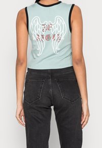 Person klädd i en ljusblå ärmlös crop top med vita änglavingar och röd text "TO ANGEL" på ryggen, tillsammans med svarta högmidjade jeans.