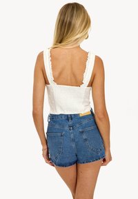 Top corto blanco texturizado con tirantes con volantes; combinado con pantalones cortos de mezclilla desgastados de cintura alta. Costuras y bolsillos visibles.