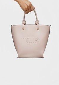 Bolsa de tote texturizada en rosa empolvado con asas anudadas y logotipo "TOUS" en relieve; acentos negros en la base; silueta redondeada, diseño amplio.