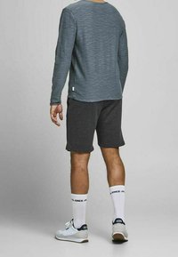 Homme portant une chemise gris à manches longues, un short gris foncé, des chaussettes blanches montantes avec le texte "JACK JONES", et des baskets blanches, debout de dos.