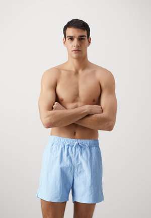 Pantalones cortos de algodón azul claro con cintura elástica y cordón, que presentan un corte holgado y bolsillos laterales con abertura. Modelo de pie con los brazos cruzados.