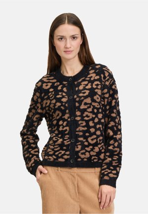 Model in een zwarte cardigan met een bruine luipaardprint, gecombineerd met lichtbruine corduroy broek, staande met handen in de zakken.
