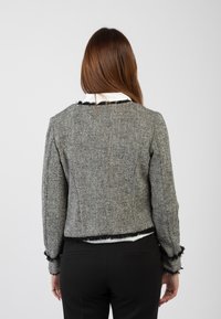 Giacca a quadri houndstooth in bianco e nero con una vestibilità cropped, bordi sfilacciati e maniche strutturate, indossata su una camicia bianca con colletto.