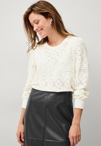Pull en tricot de couleur crème avec un motif floral texturé, à manches longues et à encolure arrondie, associé à une jupe en cuir noir ajustée.