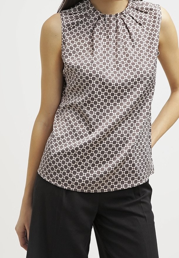 Femme portant un haut sans manches marron et blanc à motifs avec un col plissé, associé à un pantalon noir ample, debout avec la main sur la hanche.