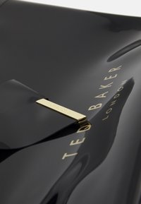 Czarna torebka z lakierowanej skóry z złotym metalowym akcentem, z klapk�ą i wytłoczonym napisem "TED BAKER LONDON". Gładka tekstura.