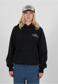 Sweat à capuche noir surdimensionné avec une poche avant, orné de l'inscription "PROHIBITED" brodée en or. Associé à un pantalon bleu et une casquette beige et noire.