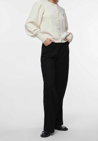 Cárdigan de punto crema con mangas abullonadas y grandes botones, combinado con pantalones de pierna ancha negros de talle alto y zapatos negros brillantes.