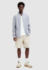 Lys blå skjorte, hvit T-skjorte under, beige shorts og kremfargede sneakers. Antrekket har en avslappet og ledig passform.