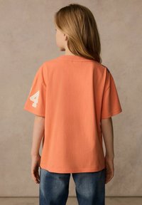 Kortärmad, korallorange t-shirt med en avslappnad passform, som har ett vitt nummer "4" och blå detaljer vid axeln. Buren med jeans.
