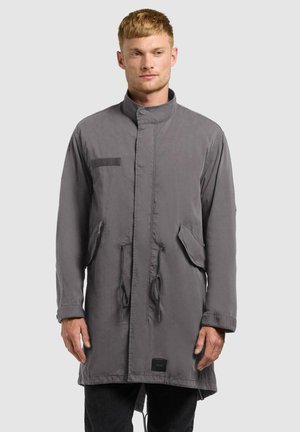 Mann trägt einen grauen langen Mantel mit Klappentaschen und Kordelzügen, steht vor einem schlichten hellen Hintergrund.