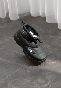 Juodas izoliuotas slip-on tipo batelis su "The North Face" logotipu šone, su raištelių uždarymu, gulintis ant pilkos plytelėmis išklotos grindų šalia baltos užuolaidos.
