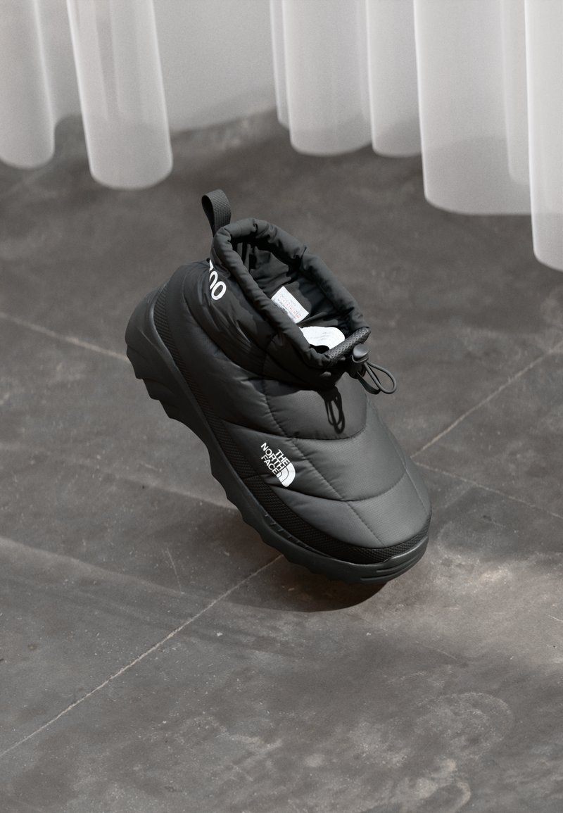 Juodas izoliuotas slip-on tipo batelis su "The North Face" logotipu šone, su raištelių uždarymu, gulintis ant pilkos plytelėmis išklotos grindų šalia baltos užuolaidos.