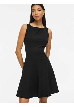 Mujer que lleva un vestido negro sin mangas hasta la rodilla con un corpiño ajustado y falda acampanada, de pie frente a un fondo blanco liso.
