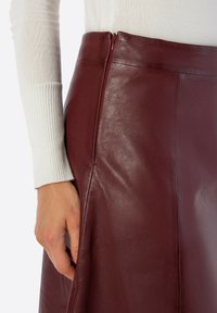 Bordeauxrode leren rok met een gladde textuur, voorzien van een onopvallende ritssluiting en een getailleerd ontwerp. Hand raakt de zijnnaad aan.