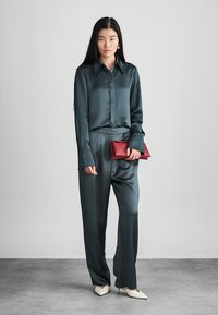 Μπλούζα από σατέν σε τιρκουάζ χρώμα με μακριά μανίκια και κουμπιά μπροστά, συνδυασμένη με παντελόνι του ιδίου χρώματος. Το μοντέλο κρατάει μια κόκκινη τσάντα και φοράει άσπρα τακούνια.