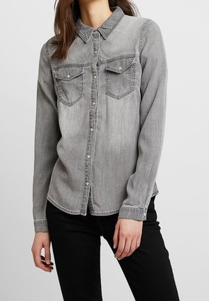 Woman wearing a faded gray button-up denim shirt with chest pockets and black pants, standing with one hand near her thigh.