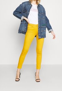 Takit denim dengan saku, kaos putih bertuliskan "EMME", dan celana skinny kuning. Sepatu hak hitam dengan tali melengkapi penampilan.