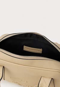 Bežs ādas rokassoma ar atvērtu rāvējslēdzēju, kurā redzams melns iekšpuse un izšūts "KARL LAGERFELD" uzlīme iekšpusē.