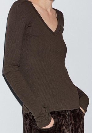 Personne portant un haut à manches longues en maille côtelée marron foncé avec un col en V, les mains dans les poches d'un pantalon texturé marron foncé, sur un fond clair.