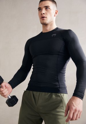 Schwarzes langärmliges Sporthemd mit taillierter Passform und dezentem Logo, kombiniert mit lockeren olivgrünen Shorts, während eine Hantel in einer Hand gehalten wird.