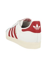 White leather sneaker with red accents, featuring three red stripes, a logo on the heel, and a textured off-white rubber sole.
