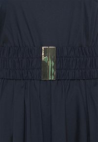 Robe en tissu noir avec taille froncée et boucle métallique rectangulaire dorée gravée de "Michael Kors" au centre.