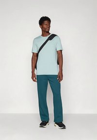 Man in een lichtblauw overhemd met korte mouwen, teal gekleurde broek, zwarte sneakers en een zwarte schoudertas, staand tegen een effen lichte achtergrond.