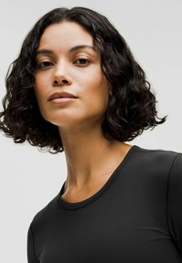 Črna rebrasta majica z dolgimi rokavi in okroglo ovratnikom. Tkanina je gladka z rahlo teksturo. Lase ima kodraste in dolge do ramen.