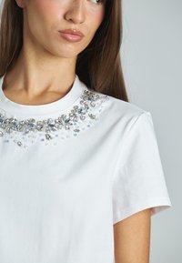 Camiseta blanca de algodón con un cuello decorado con cuentas que incluye gemas falsas transparentes y azules, mangas cortas y un diseño clásico de cuello redondo.