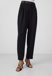 Zwarte geplooide broeken met een getailleerde pasvorm, voorzien van een gladde tailleband en gestructureerde pijpen. G gedragen met zwarte sandalen met hakken en bandjes.