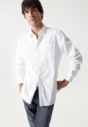 Camisa blanca de manga larga con cuello abotonado, un bolsillo en el pecho y dobladillo recto, combinada con jeans de mezclilla azul oscuro.